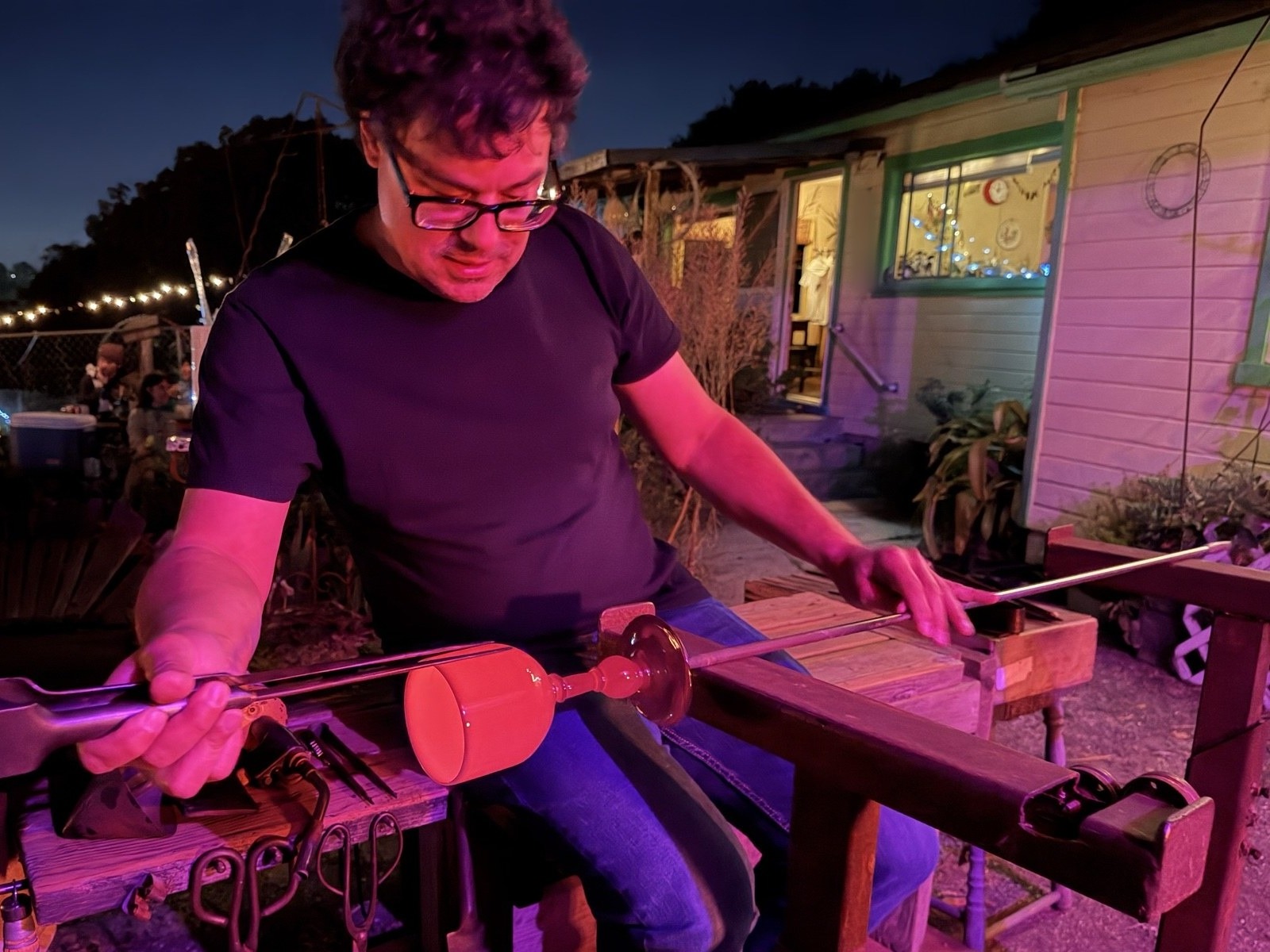 Arlo Fishman at the glassblowing bench at dusk, shaping a red goblet on the pipe